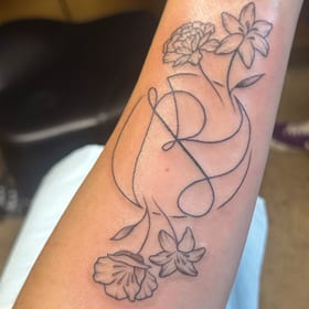 Fine-line forearm tattoo of the name Amber stacked into one flowing line, with lilies and a carnation